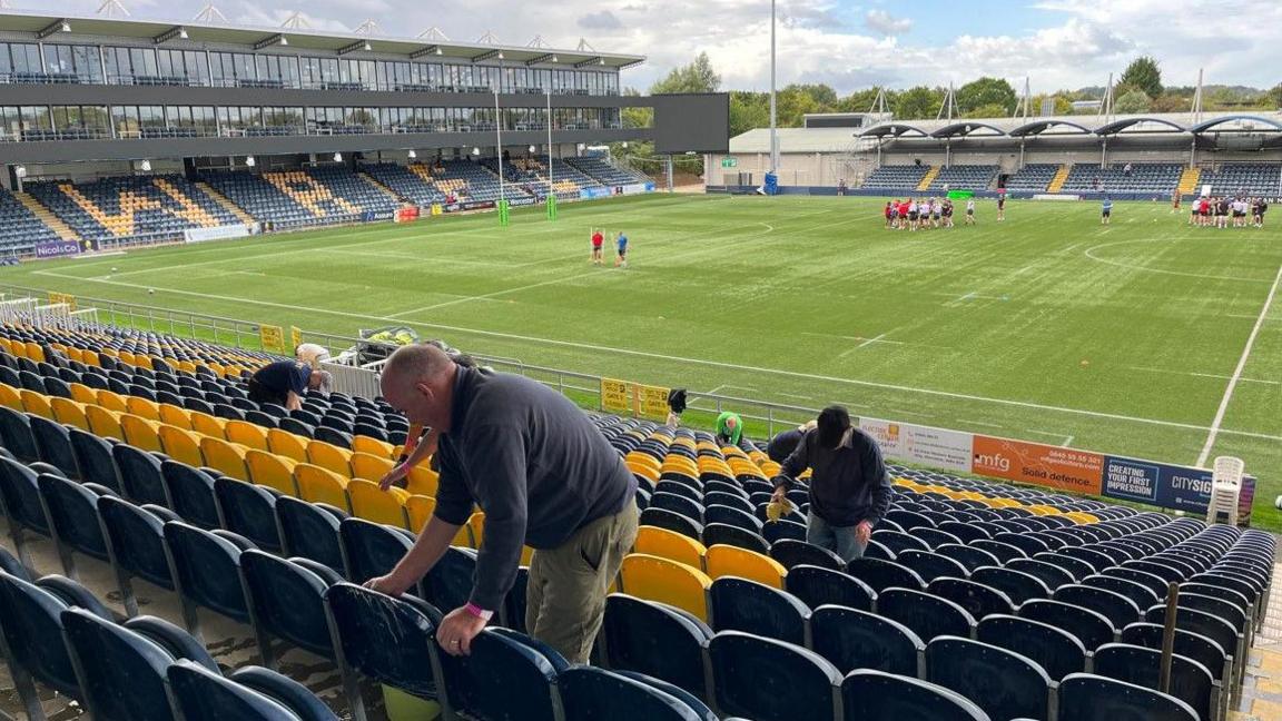 A man in the foreground and others are cleaning seats. The photographer in the same stand is not near ground level and the image includes two other stands - to the left and opposite the stand they are in. Some people are on the pitch in two groups.