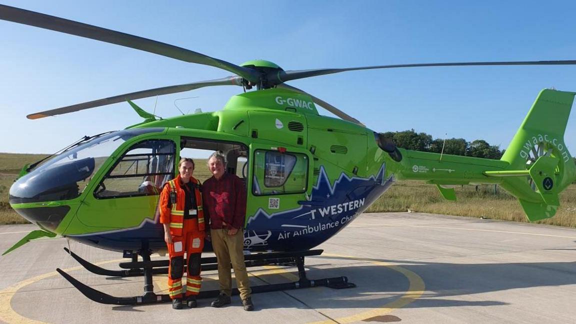 Mark standing with a member of the Great Western Air Ambulance Charity in front of a green helicopter on a helipad.