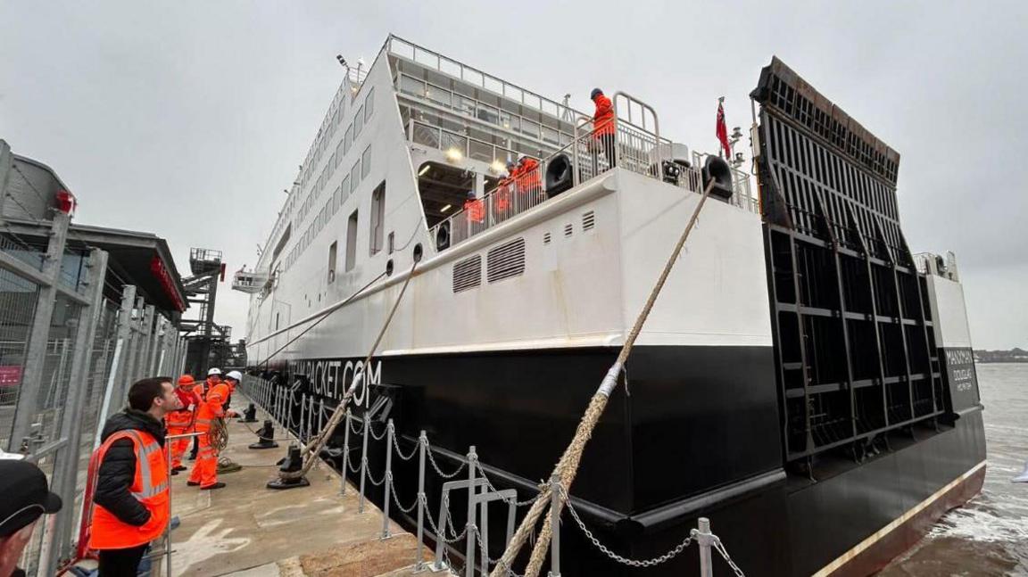 The stern of the Manxman ferry tied up next tot he dock in Liverpool with men dressing in bright orange high visibility jackets on the boat and on the dockside.