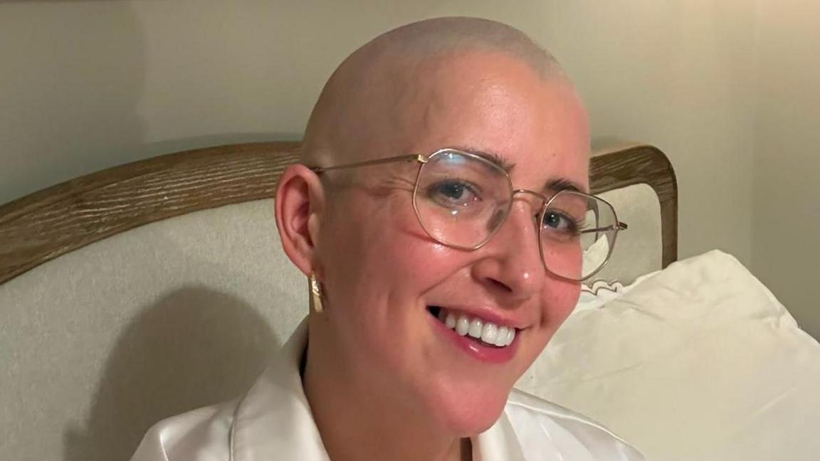 Kim Borthwick is sitting up in bed in a house. She is wearing white, silky pyjamas and large glasses. She has no hair and is smiling at the camera