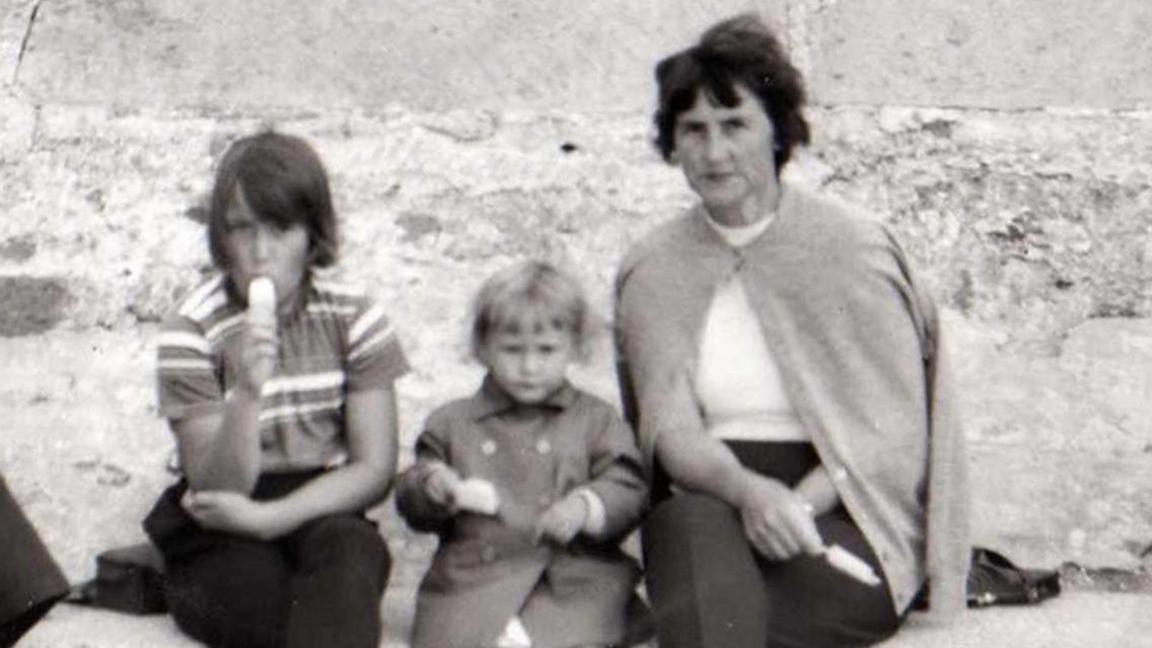 A black and white image of a mother with two children sitting on rocks by the sea. All three are eating ice lollies. The girl on the left is around 8 and wearing a striped top and black trousers. A 3-year-old girl in the middle wears a double breasted coat and her mother has a cardigan draped over a white top and black trousers.
