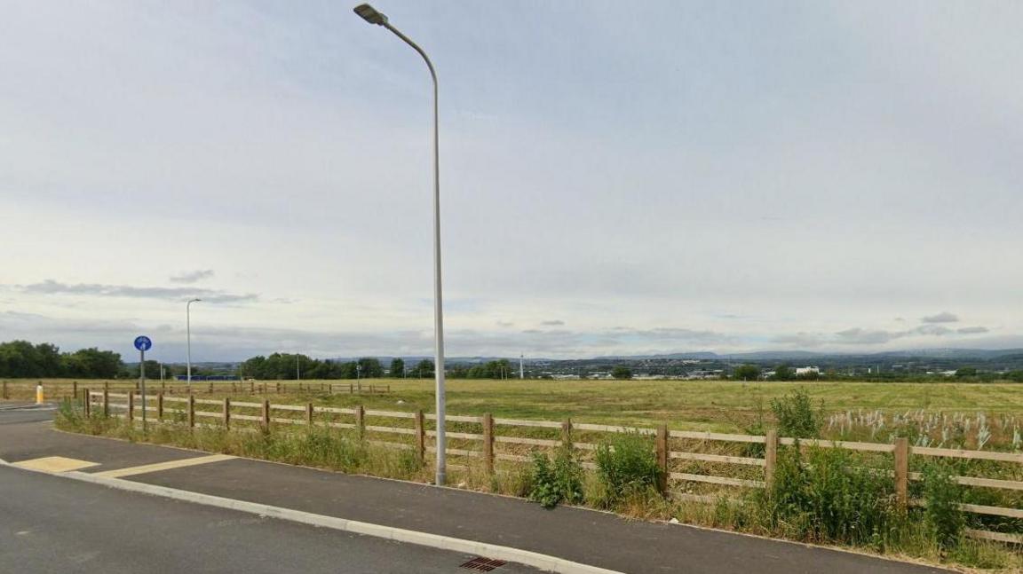 A screenshot of a Google Map street view. It shows a street with a pavement beside an empty field, enclosed by a wooden fence.