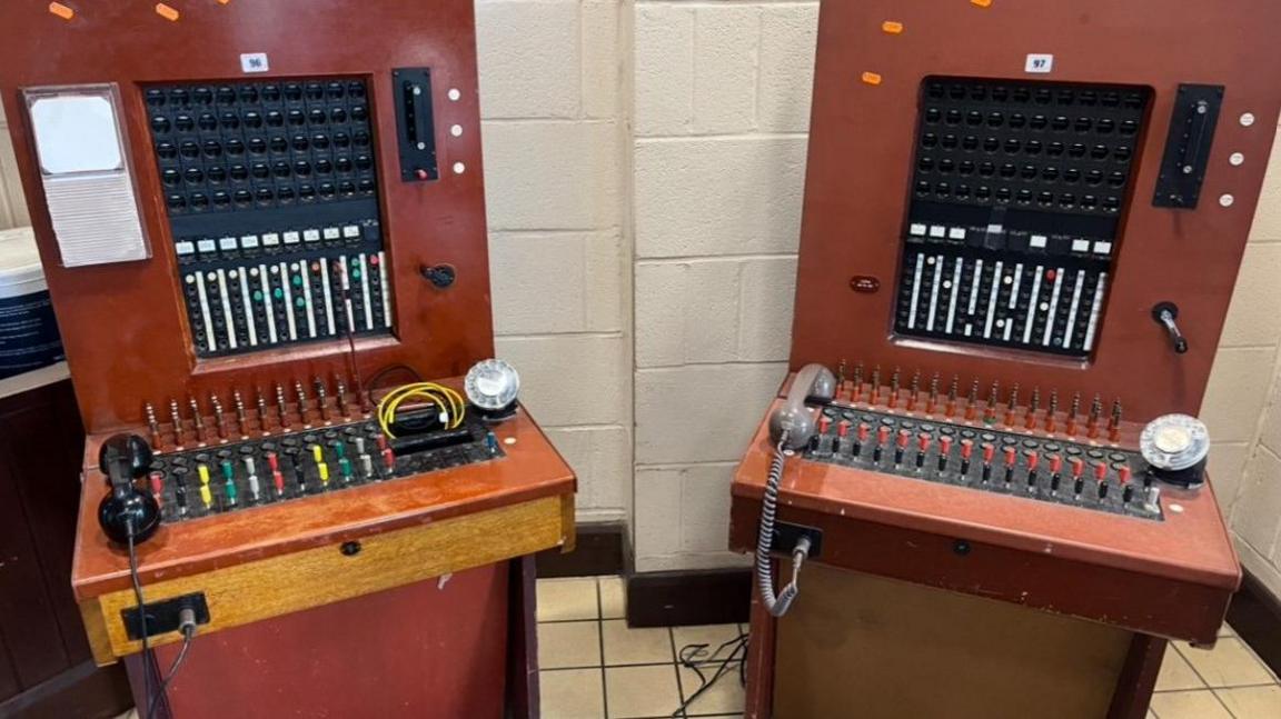 Two exchanges are pictured, with both primarily vertical, with a horizontal section containing at least a dozen buttons and a telephone receiver.