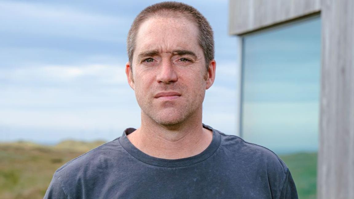 Martin Dickie, who has dark cropped hair, standing staring at the camera with a serious expression on his face. A field and a grey wooden building with a glass window can be see in the background. He is wearing a dark blue t-shirt.