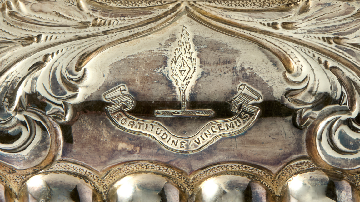 Close up of the crest on the silver tea set. The words have been engraved onto a ribbon like shape with a shape like a diamond on fire above it.