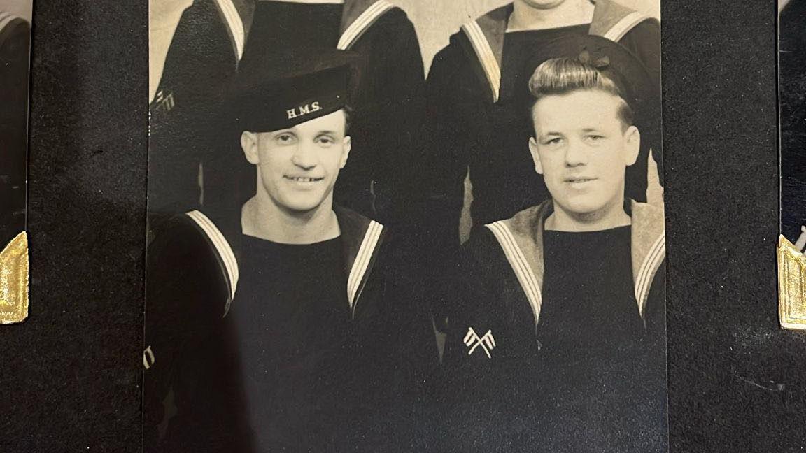 Black-and-white photo of two sailors in the Royal Navy. They are wearing uniforms. The man on the left is wearing a hat which reads 'HMS'. 