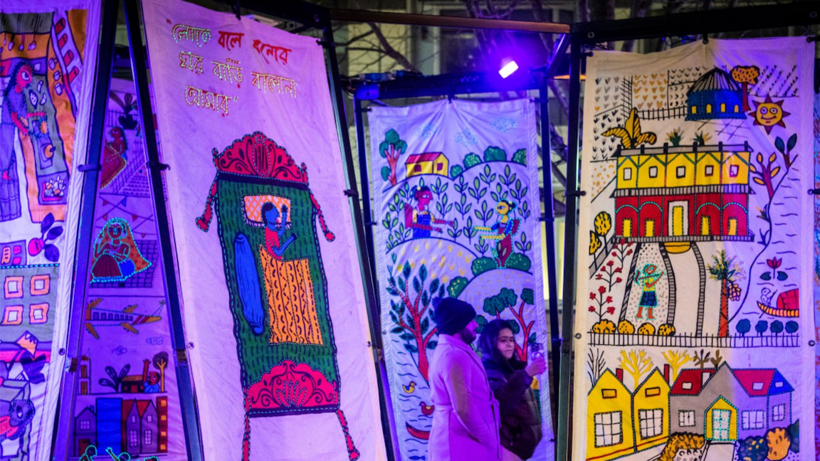 A close up view of some of the tapestries. One in the foreground of the picture displays a scene with a person lying in a be, and is painted in bright colours, including green, yellow, blue and red. Another tapestry that is prominent in the image displays a scene with various buildings and also greenery. This second tapestry consists of bright yellows, reds and blues.