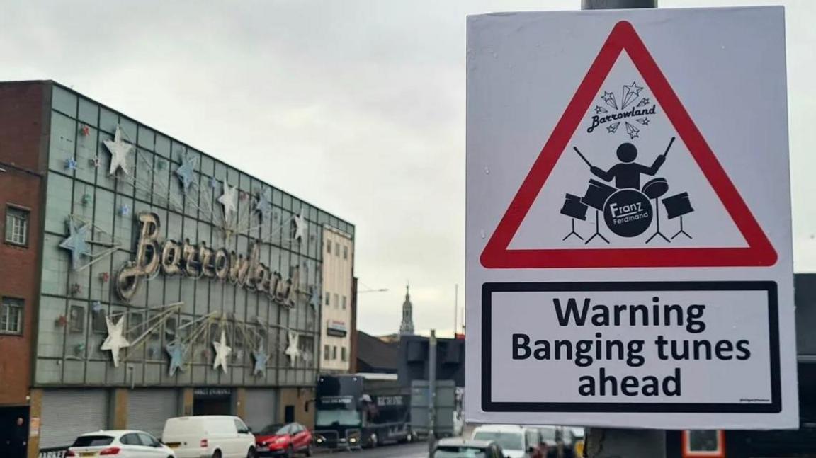 A red triangle warning sign that reads "Warning banging tunes ahead" with a cartoon man playing a drum set. The sign is in front of Glasgow's Barowland sign.