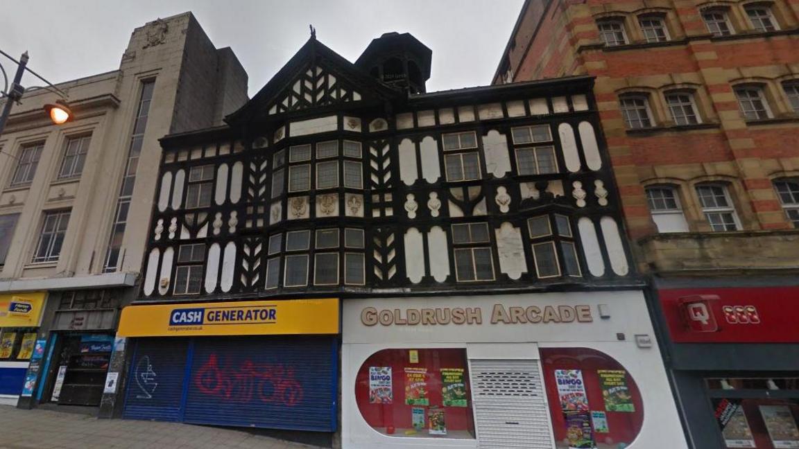 A black-and-white mock-Tudor building, with ornate black and white woodwork, windows and a bell tower. On the ground floor is a "Cash Generator" shop with a yellow sign and blue shutters, and a "Goldrush Arcade", with a white sign, white shutters and red windows.