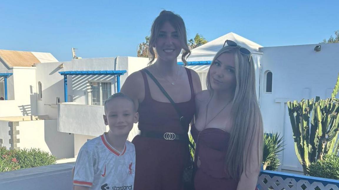 A woman with brown hair in a bun, wearing a maroon jumpsuit with a black and gold belt and black cross-body bag. She has one arm around a young blonde boy in a red and white football shirt, and the other arm around a teenage girl with long blonde hair, wearing a maroon co-ord outfit and sunglasses on her head. The trio are stood on a white balcony with white-wash buildings and greenery behind them. They are all smiling and looking at the camera.