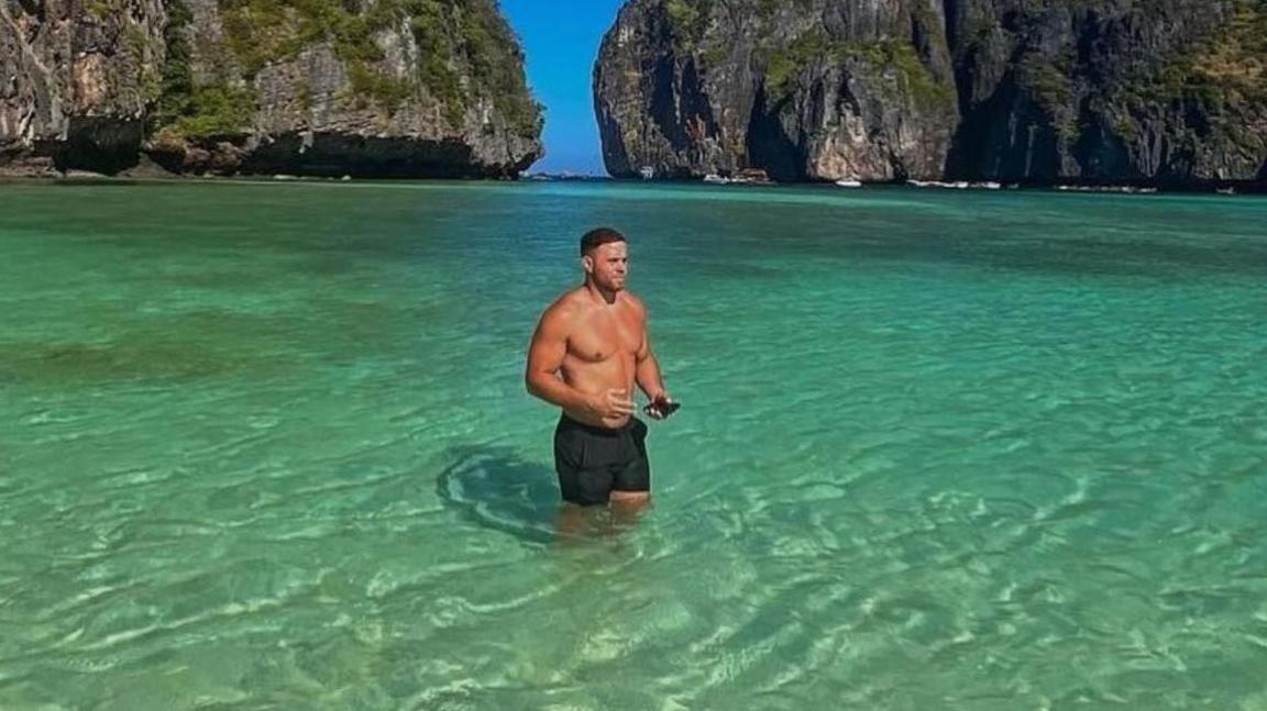 Corey Beavis pictured in black swim shorts standing in clear blue green water with mountains behind