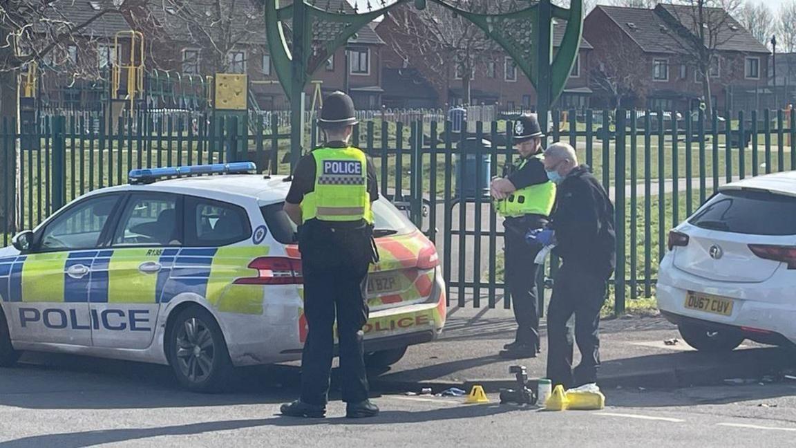 Three police officers stood near evidence markers and a police car