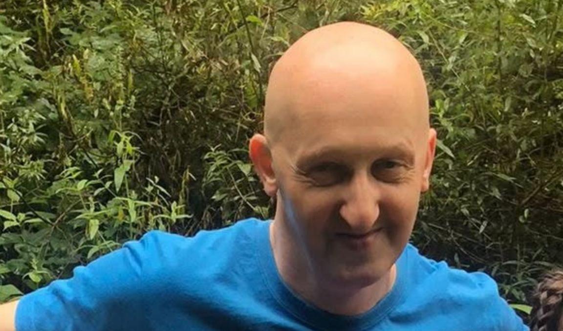 A man smiles at the camera. He is bald and is wearing a blue t-shirt. There is green foliage behind him.