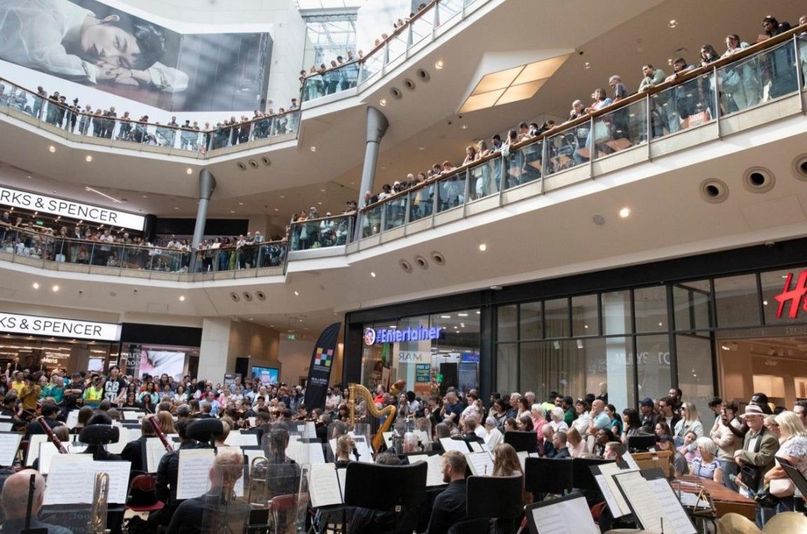 A full orchestra on the bottom floor of a shopping centre, large crowds are gathered and watching from the floors above