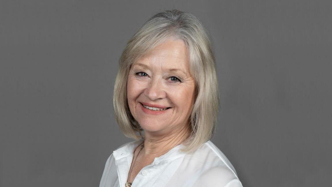 A head shot of Dame Yve Buckland who is wearing a white shirt with a grey background