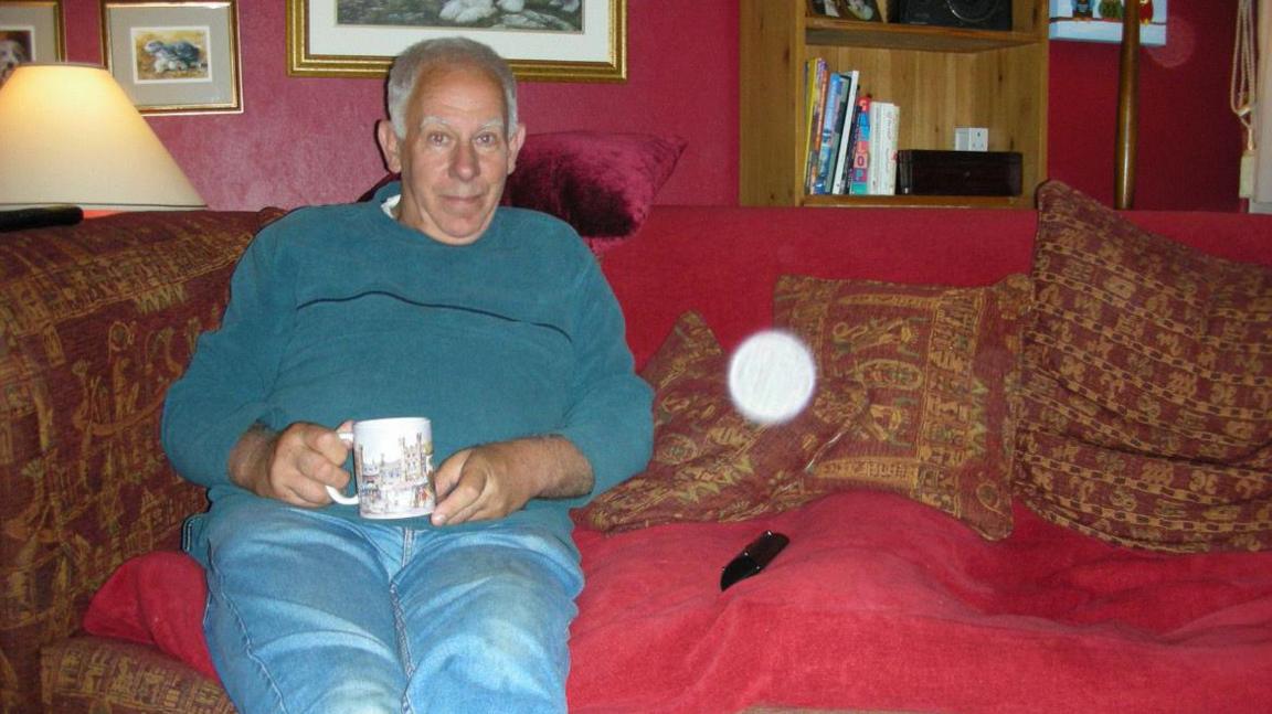 Dave Price is sitting on a large red sofa with red cushions. Behind him is a lamp which is turned on. He is holding a mug. Next to him is a white spot on the photograph, which he and Liz Price say is a light anomaly