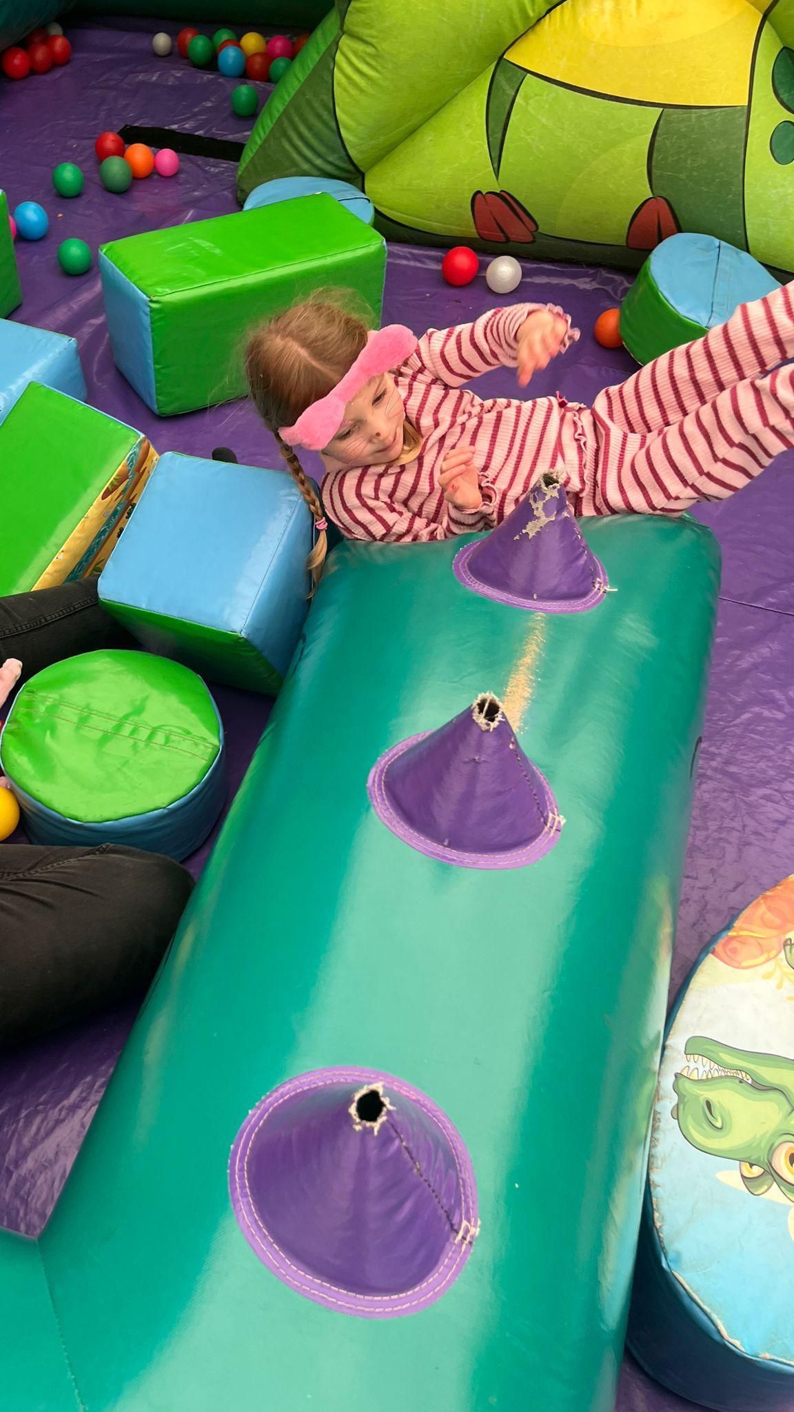 A young girl is lying down on a purple sheet-type covering on the floor. There are some small foam blocks around her and what looks like an inflatable green low wall with purple cones with holes in the top. Part of a woman holding a baby can be seen sitting down on the floor nearby.