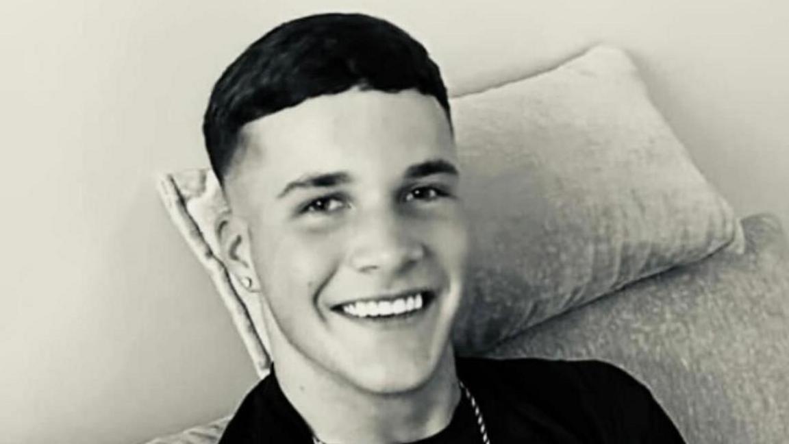 A black and white photo of a young man with short dark hair, sitting on a sofa and smiling at the camera.