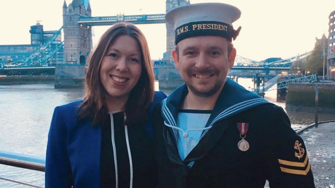 Mr Oliver and his wife Emily. Mr Oliver is in a Naval uniform. They stand in front of Tower Bridge in London.