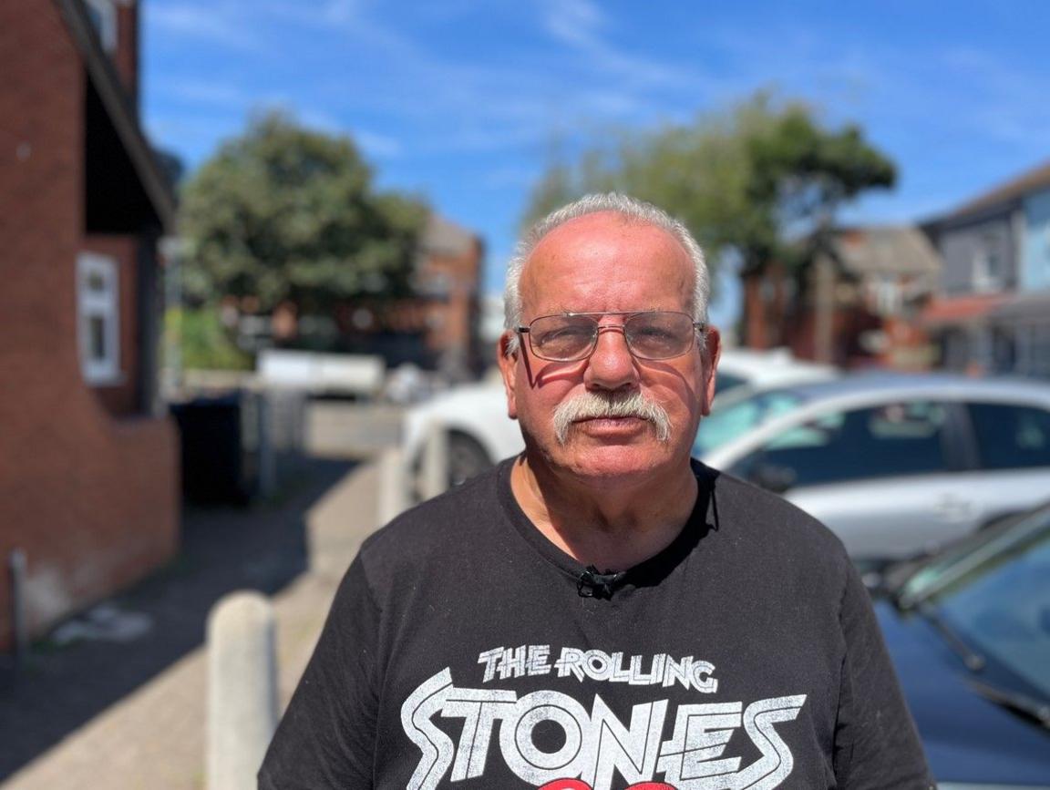 A man with a black T-shirt with the words The Rolling Stones in white is wearing glasses and has a moustache. Properties and cars are in the background.