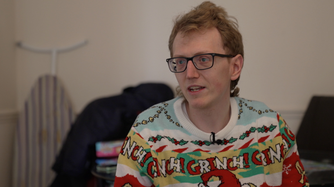 Sam is wearing a bright and colourful jumper and dark-framed glasses. 