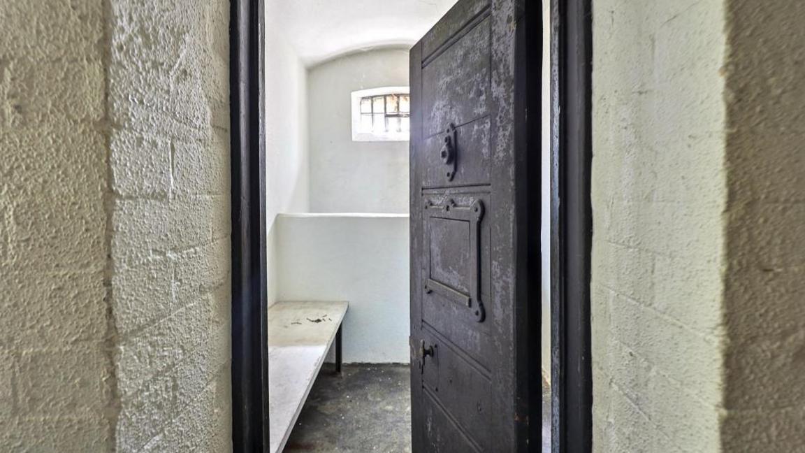 An imposing heavy grey metal cell door stands ajar, a bed can just be seen inside.