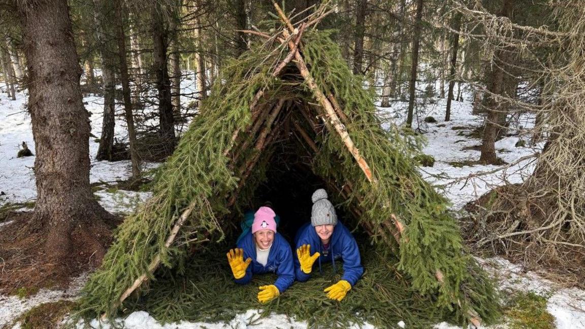 Two people in blue coats are on the ground, both with yellow gloves and their right hand waving at the camera. One wears a pink hat, the other a grey one. They are in a triangular shelter with branches on it, in a forest with snow on the ground.