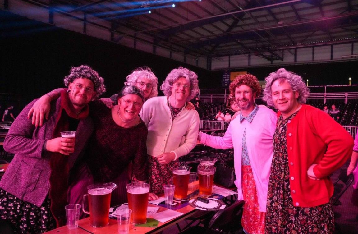 Six people are standing up by a table, which includes a number of pint glasses.