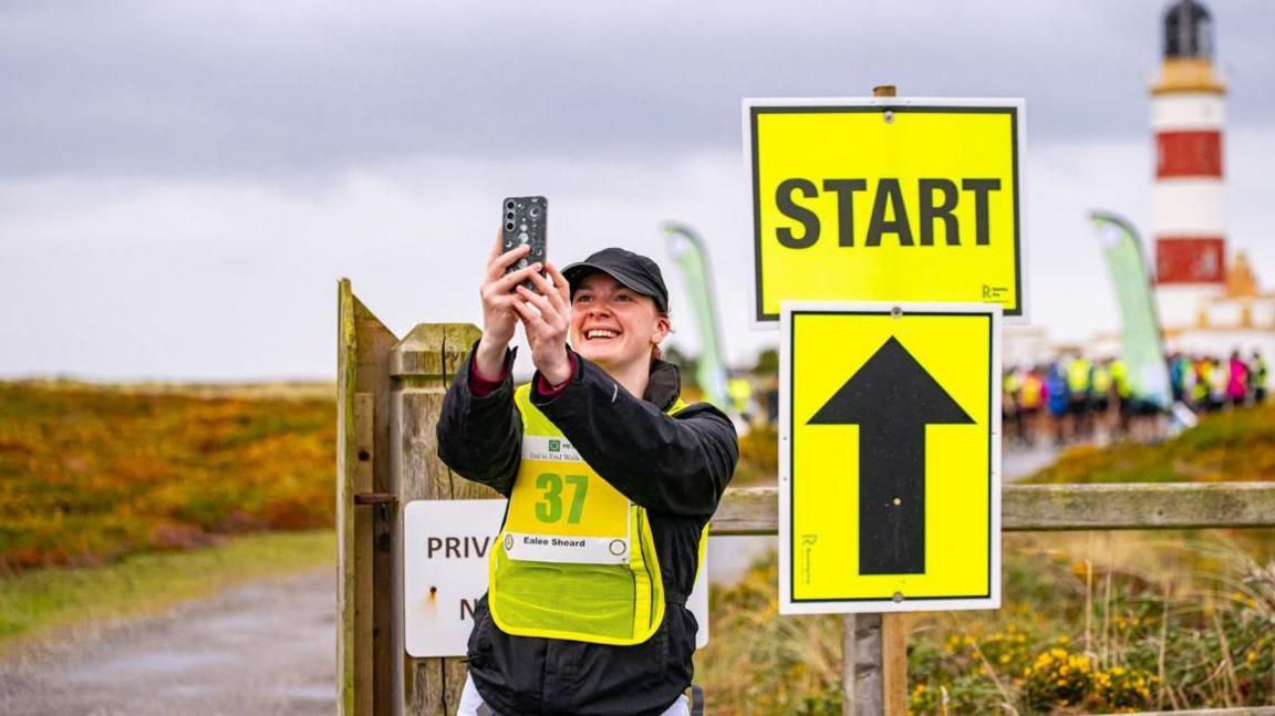 A smiling woman holding up a phone taking a selfie next to a fluorescent start sign that has a large black arrow on it. She is wearing a hi-vis tabard and a black cap and top. You can make out the red and white lighthouse at Point of Ayre and a group of walkers in the background.