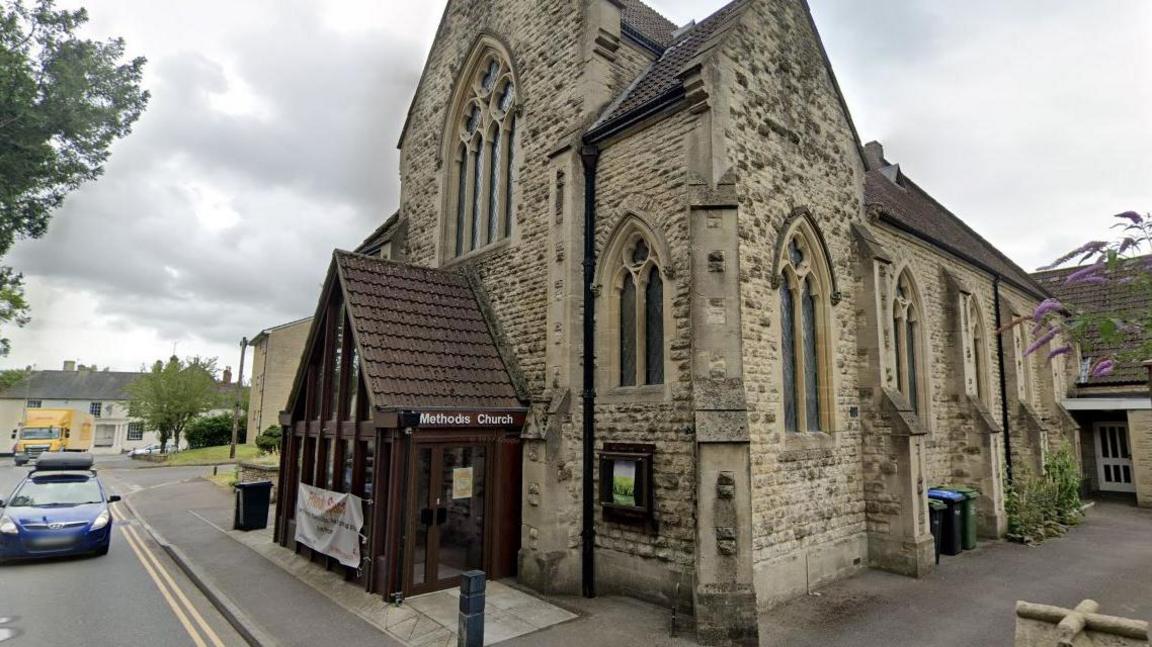 A church building beside a road. The stone church has arched windows and a small, more modern, porch attached to the front with methodist church written on it