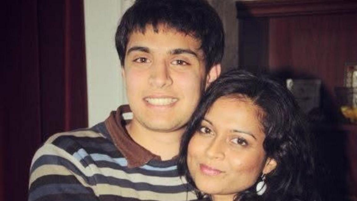 A black-haired boy with a stripy jumpter and brown polo neck t-shirt is smiling while embracing a smiling lady with black hair