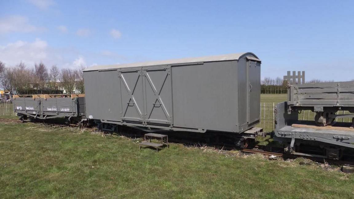 A grey 1917 emergency ambulance van with double doors sits on a light-gauge railway track between smaller supply wagons. A small step offers access to a grassy field