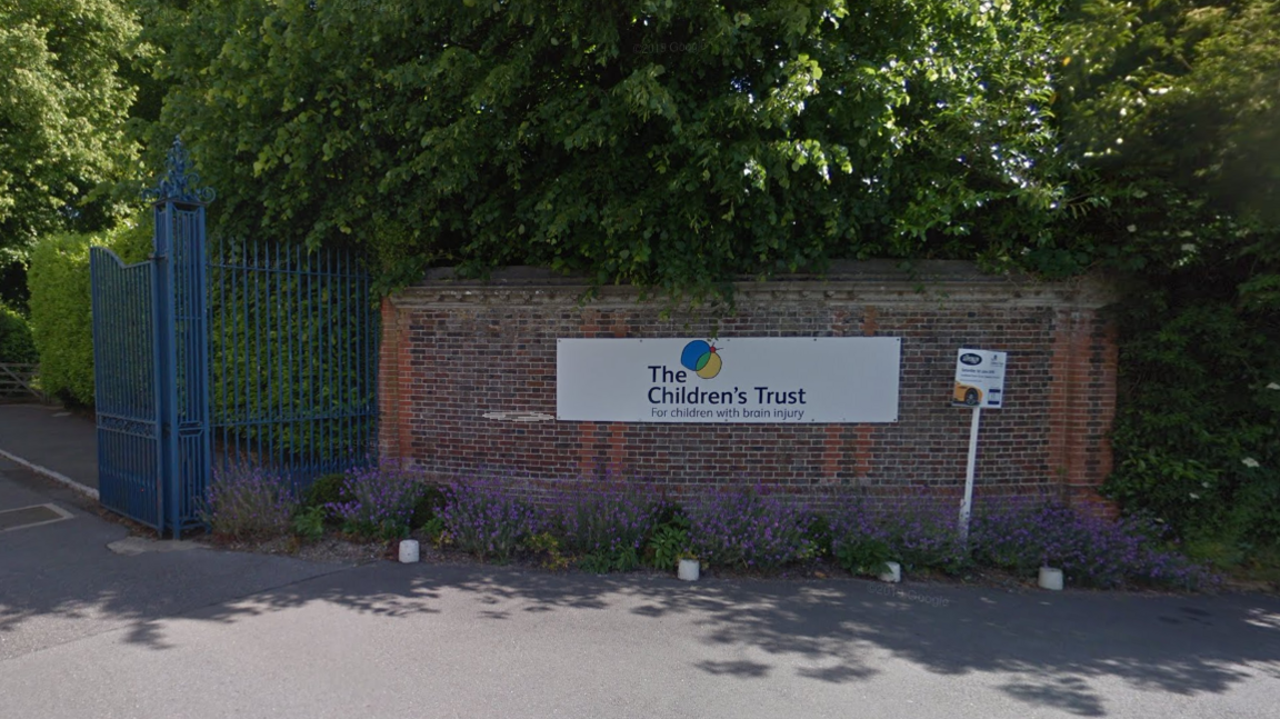 A red brick wall with a sign reading The Children's Trust adjacent to blue iron gates