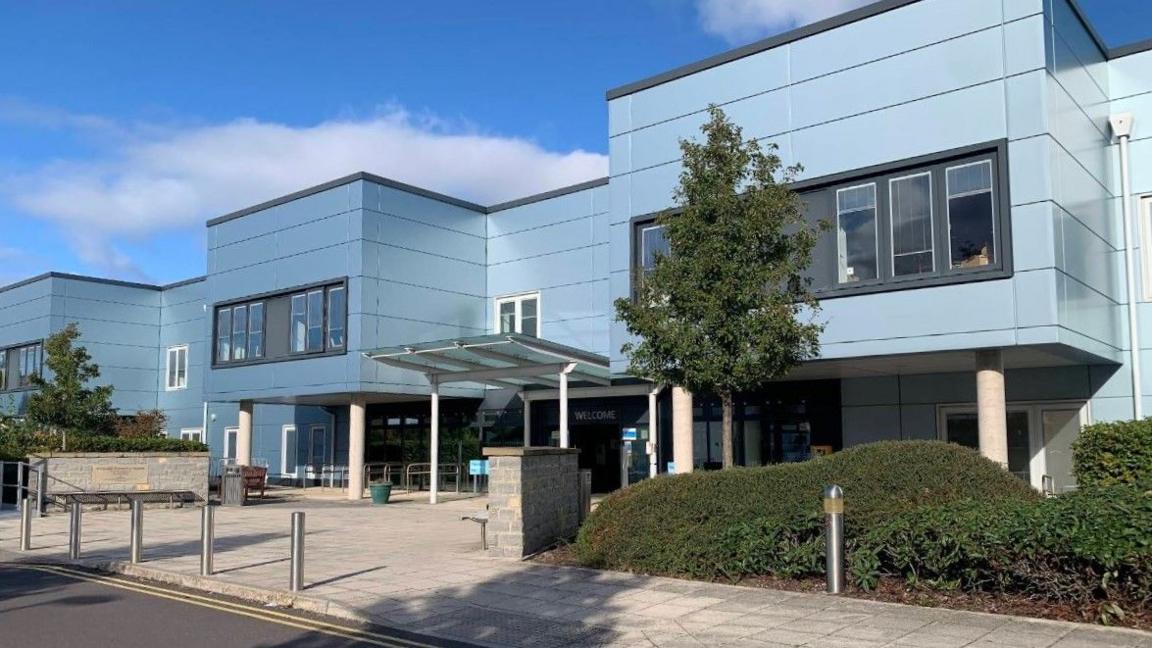 The exterior entrance to Bridgwater Community Hospital. It is a large rectangular building clad in blue metal sheets with an overhead shelter near the front.