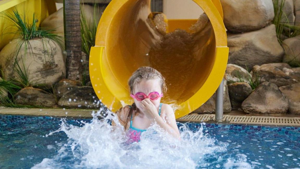 Girl wearing pink goggles entering the water after coming down a yellow slide