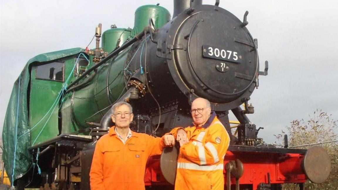 John Rice and Barry Joplin posing in front of steam locomotive 30075. It is painted green and black. They are wearing orange overalls.