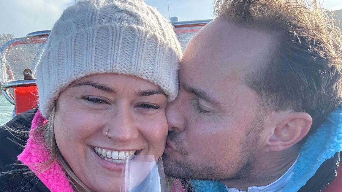 Ms Francis and Mr Tite on a boat after the marriage proposal. Mr Tite is kissing Mr Francis' cheek. She is wearing a cream bobble hat and is holding up a glass of champagne. 