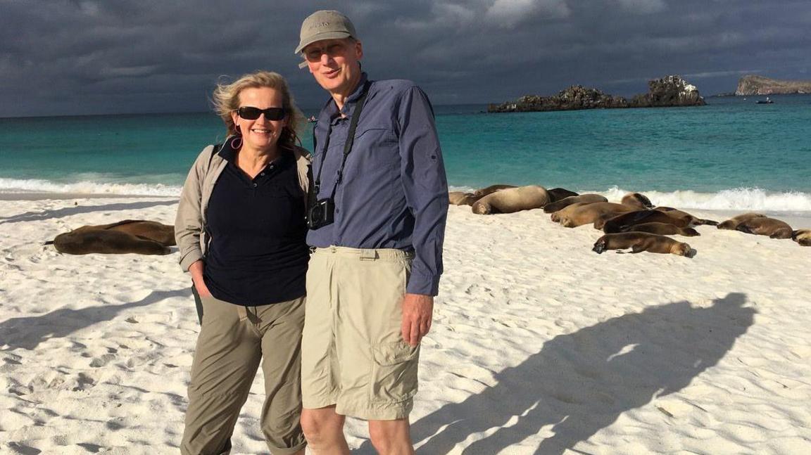 Prof Crellin and his wife on a beach with seals in the background - both smiling and wearing summer clothes and sunglasses