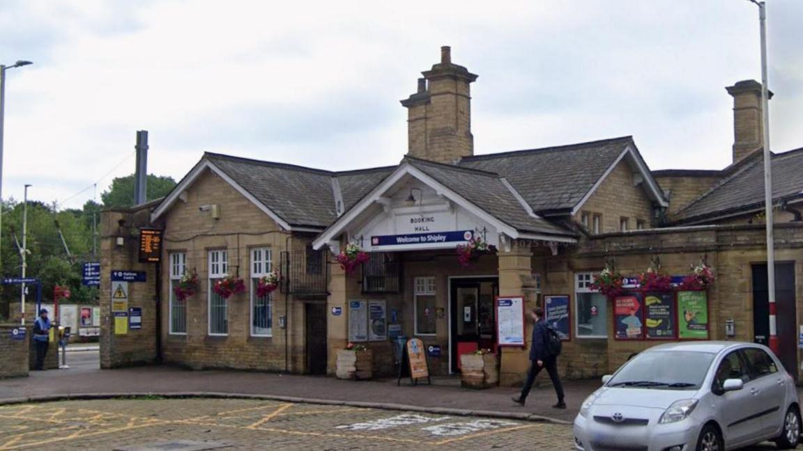 A single storey railway station building with billboard signs on the walls outside, a sign saying Welcome to Shipley above the main door and hanging baskets of flowers across the front of the building. A silver car is parked at the front of the building. A man in dark clothes with a backpack is walking past the entrance. A black sign with organe lit up live train times is outside the front of the building.
