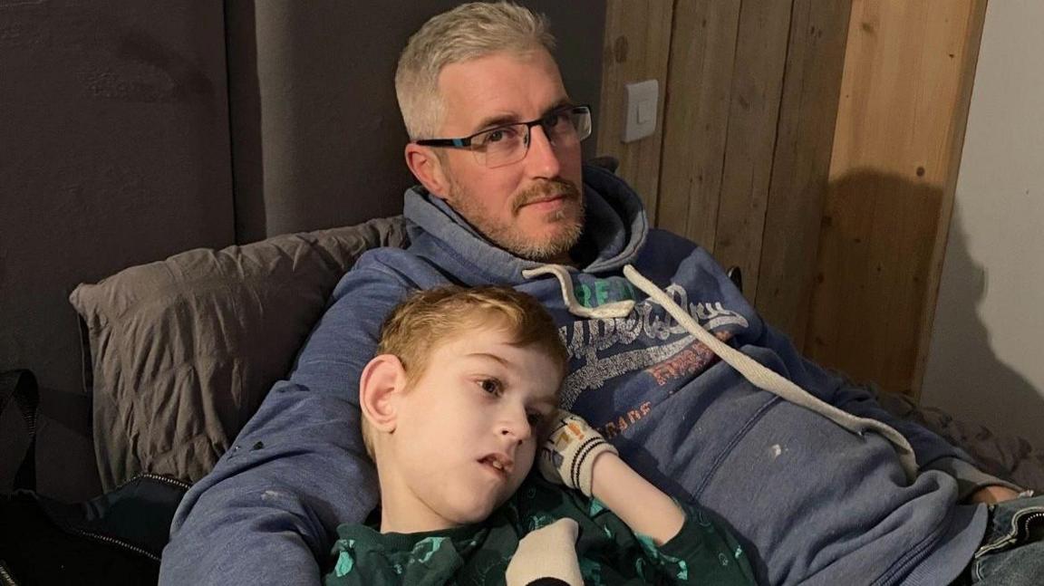 Shaun smiles at the camera with his arm around Toby. Shaun is wearing a blue hoodie and glasses.