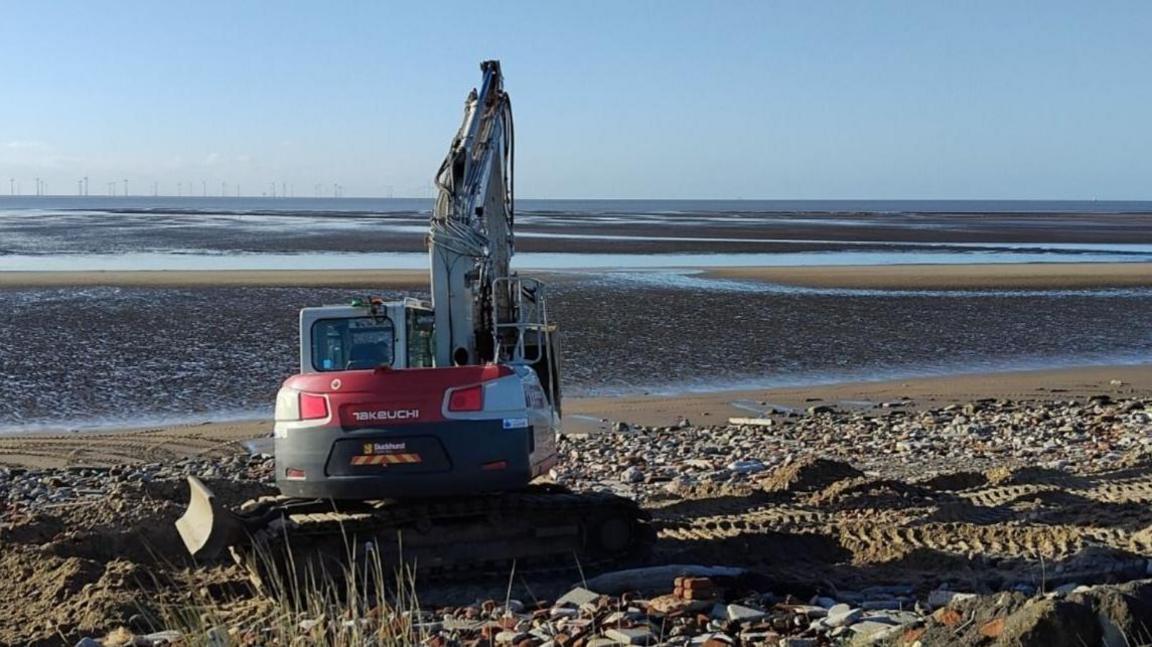 A small digger sits on a sandy beach surrounded by piles of rubble. The tide is far out into the distance, and a line of wind turbines are visible on the horizon.
