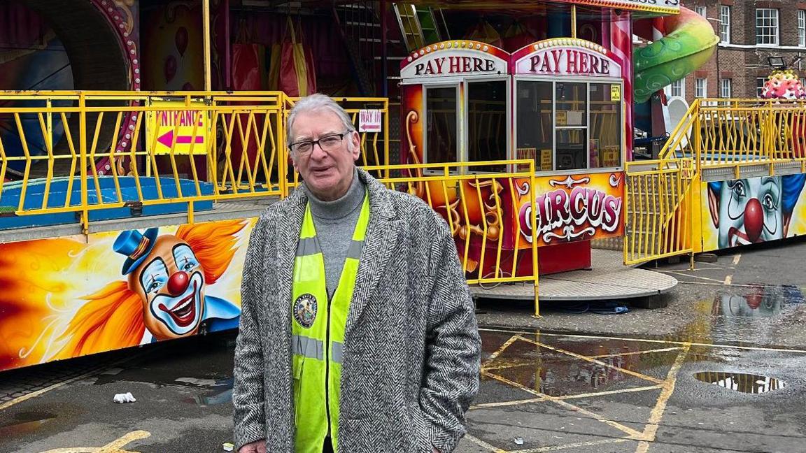 Showman Nipper Appleton stands in front of the fun house which is brightly coloured and painted with clowns. He wears a black and white wool coat with a high vis vest under it. He wears glasses and has one hand in his pocket. 