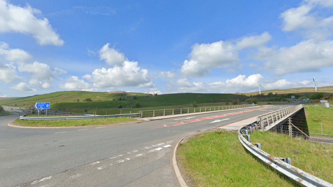 Junction 37 of the A684. It is a two-lane road with a blue sign straightway  reading 'The South M6'. There are cars in the distance and two wind turbines on the hills to the right. 