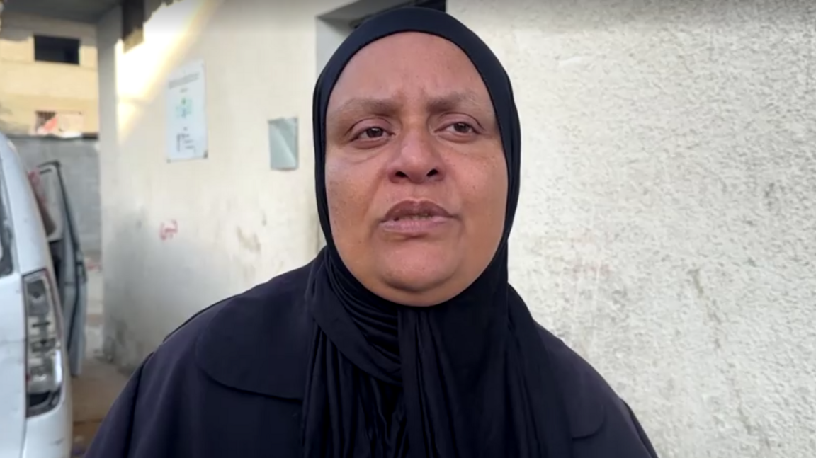 Umm Mohammed Abou Aisha, wearing black garment and head covering, with white walls and a vehicle behind