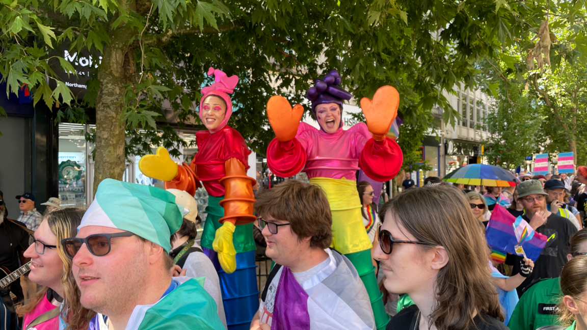 Two people can been seen towering above the crowd on stilts outside. They are dressed in rainbow costumes.