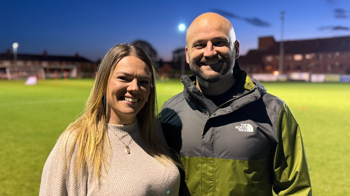 A woman wearing a light pink jumper and a gold necklace stands next to her partner who is wearing a black and green jacket which says The North Face. They are standing on a football pitch which has been illuminated as the sun has nearly set.