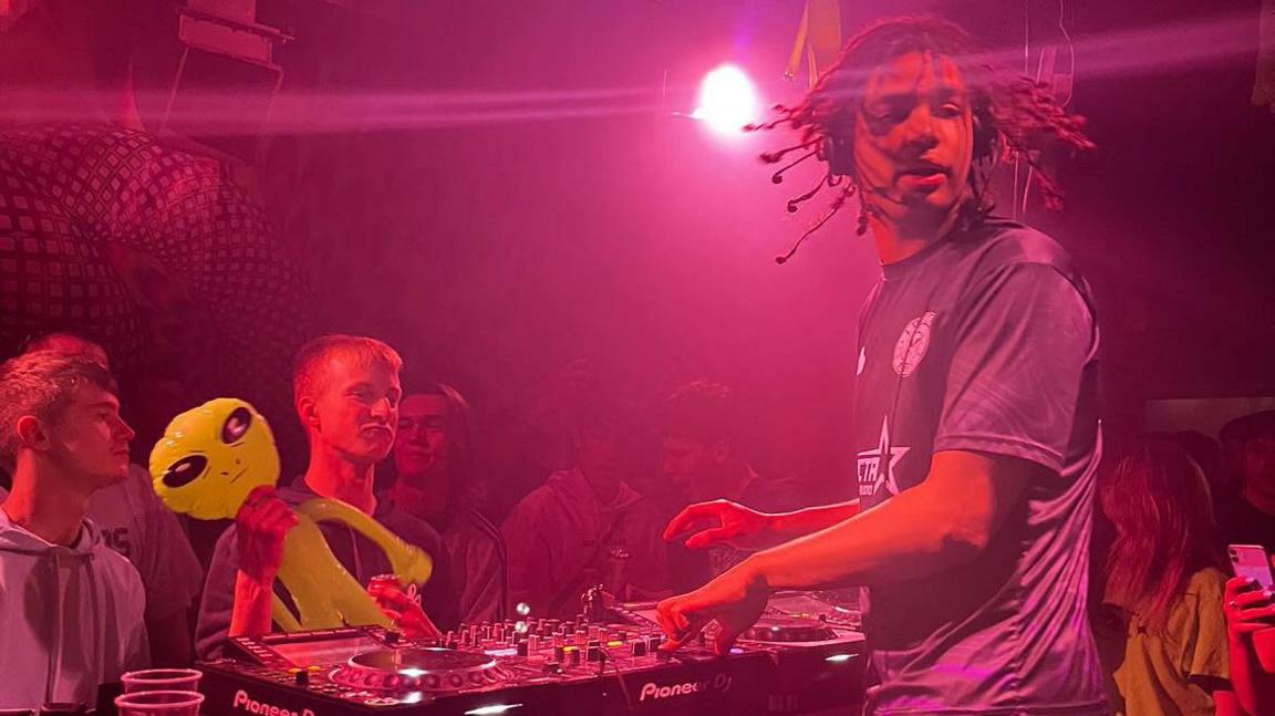 A DJ is playing a set in a nightclub which has a bright, purple light shining on the dancefloor. Anton Bailey is standing behind the turntable and looking over his shoulder. In front of him are some people watching him, one is holding an inflatable alien.