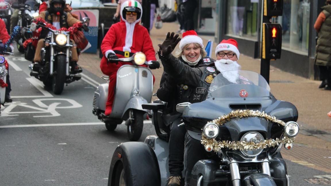 The bikers lining up the streets, dressed in Santa clothes. Their bikes are adorned with Christmas toys and tinsel. The biker in front  is waving. A woman riding behind him is smiling.