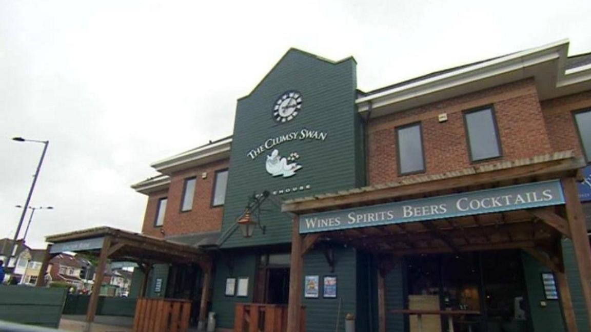 A red brick pub with green weather boards along the middle of its frontage. There is a sign that says The Clumsy Swan and another reads: Wines Spirits Beers Cocktails