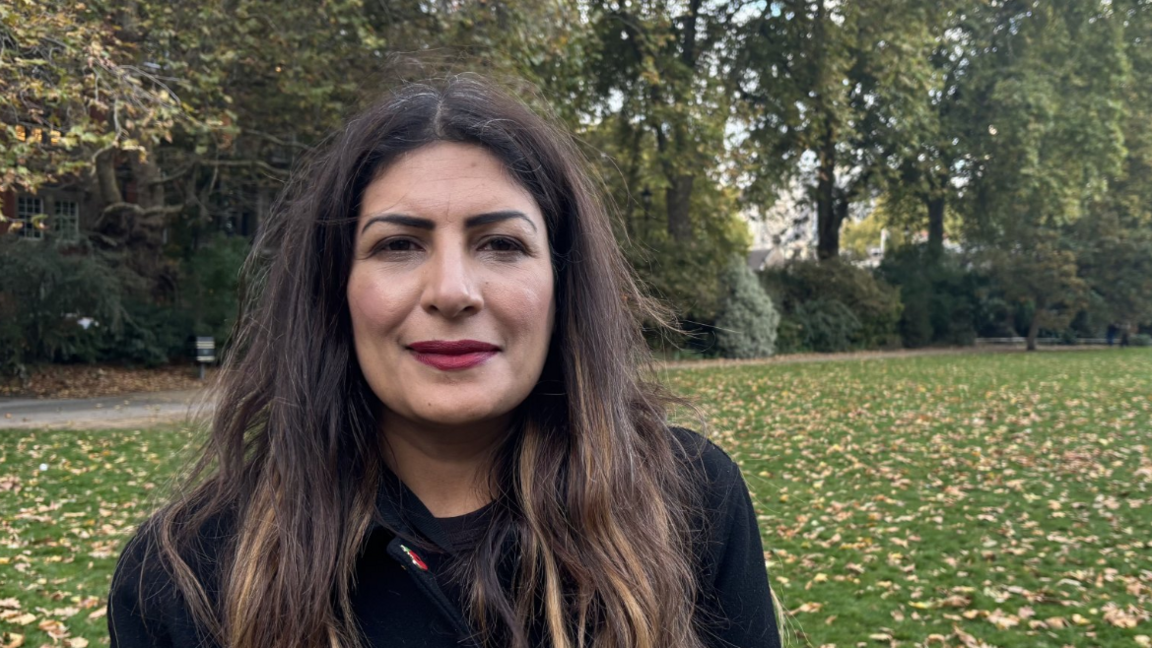 A woman with brown hair, wearing a black coat and red lipstick, smiles at the camera. Lots of trees and grass are visible behind her.